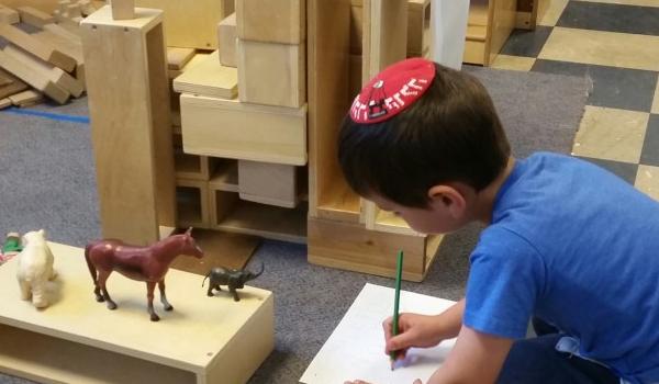 Small boy wearing a kippah sits on the floor drawing on a paper while looking at plastic animals on a block in front of him