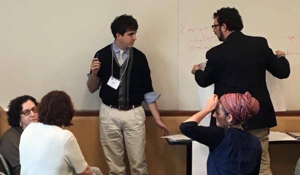 Professor Ben Jacobs writes on a large whiteboard during meeting, one other professional stands next to him, three others seated in discussion