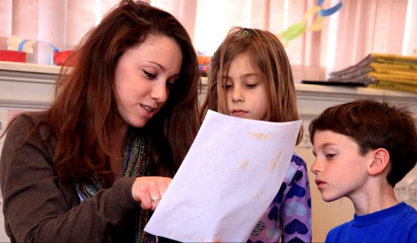 Teacher holds paper and points, two children look.