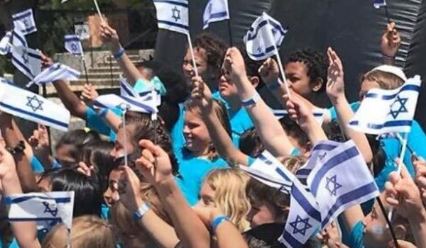 large group of children hold Israeli flags in the air at some type of rally