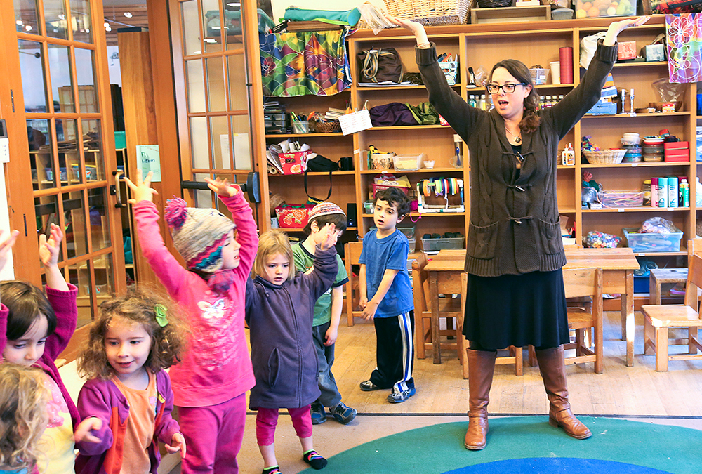 preschool teacher stands with arms raised high as preschool students by her follow her example