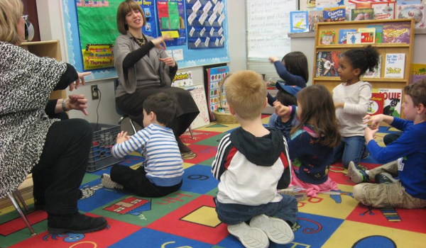 Two preschool teachers sit in chairs instructing young students sitting on a brightly colored carpet