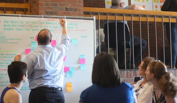 Person stands at a white board with colorful post it notes on the board. Other people are seated behind.