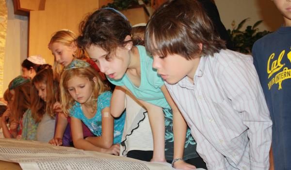 Young children lean down to look at a Torah spread across a table