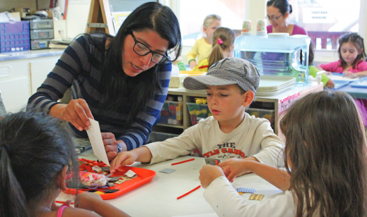 Jewish Day School teachers holds paper up for elementary student seated at table reaching for a red tray of supplies