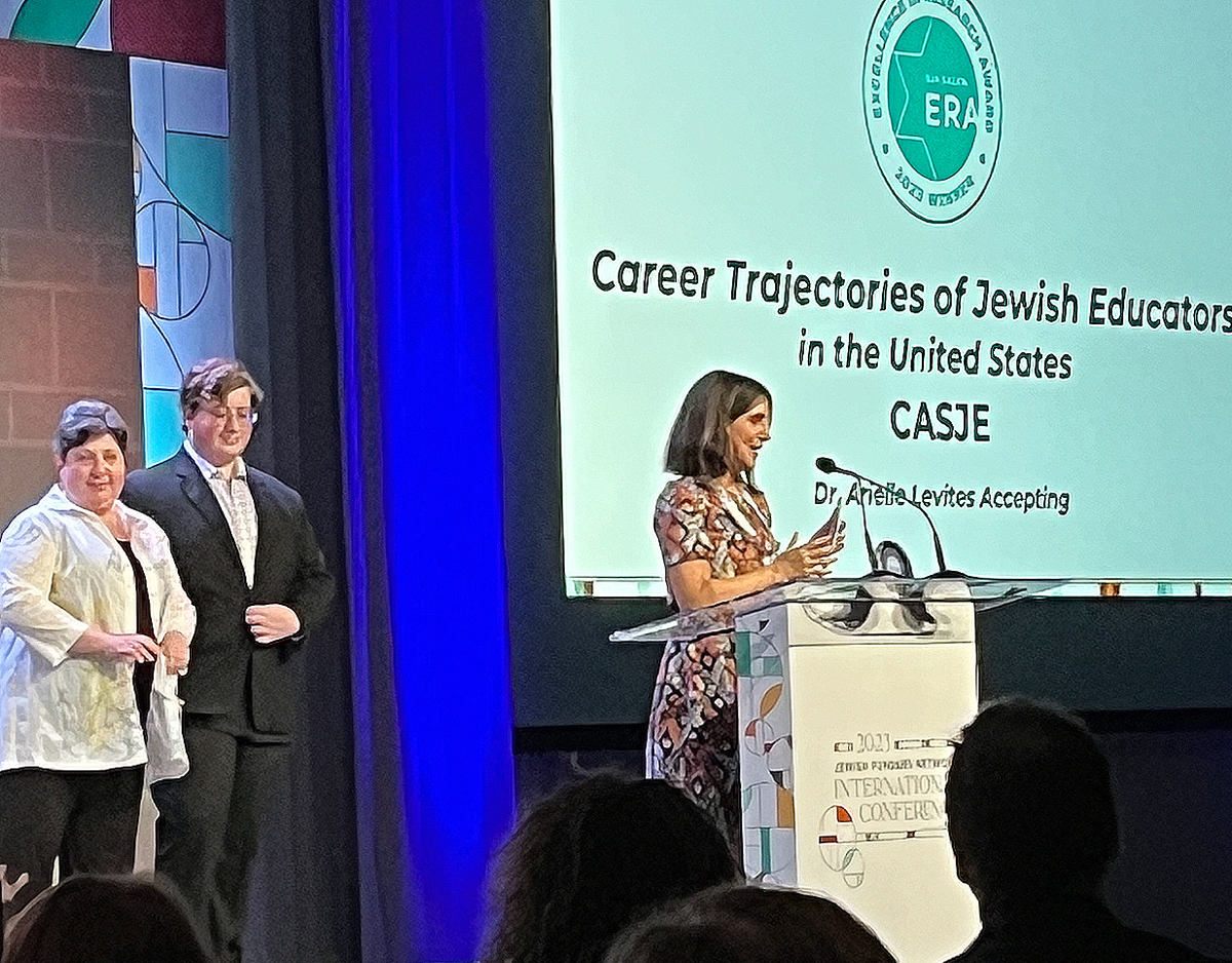 Dr. Arielle Levites stands on a podium. The Salita family stands behind her. A projector screen behind Dr. Levites reads, Career Trajectories of Jewish Education in the United States"
