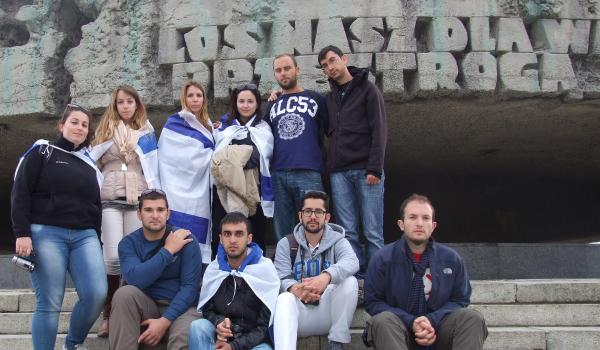 Group of people, some in Israeli flags, stand in front of a rock wall with letters.