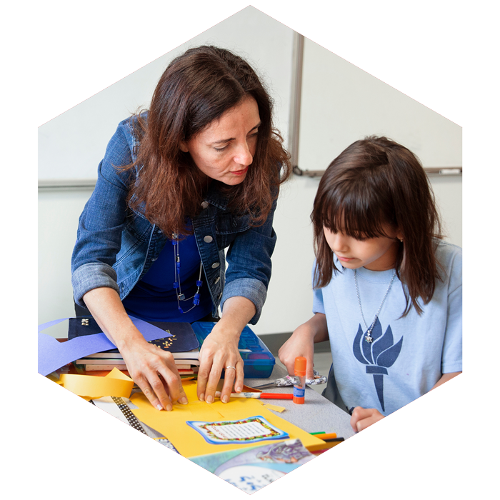Jewish Day School teacher assists elementary aged student with craft (image is hexagon shaped)