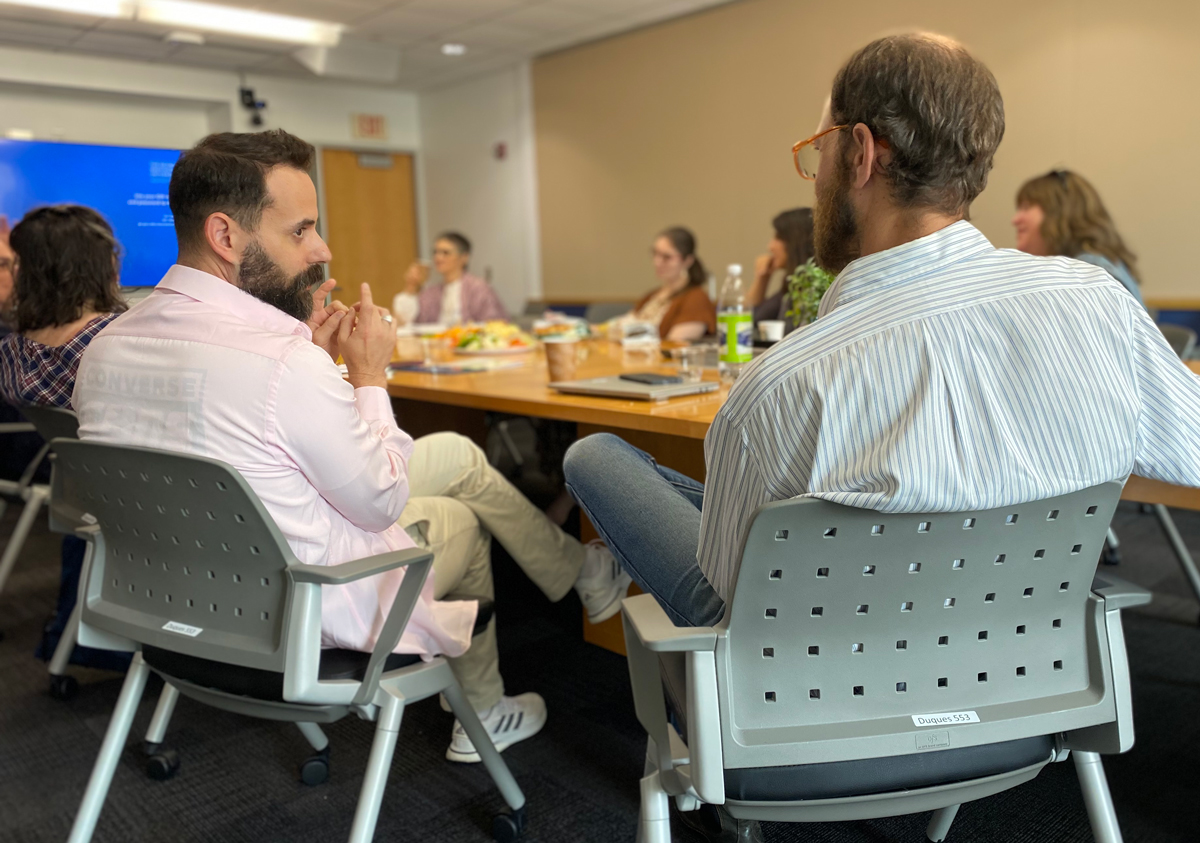 A fellow and mentor speak together with their backs facing the camera, other fellows are seated around the table in the background