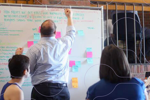 professional male writes on large white board with post-it notes scattered across it, small group watches