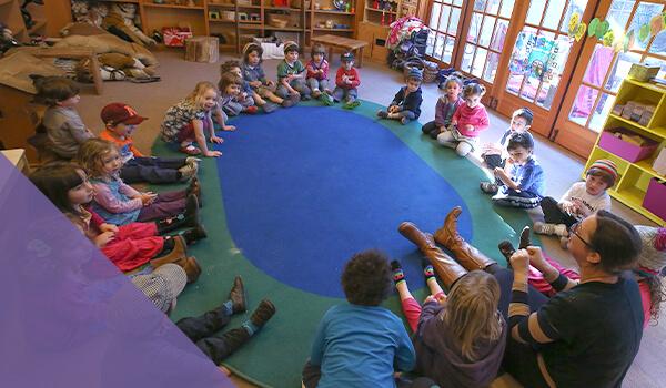 preschool students sit around round blue rug in classroom