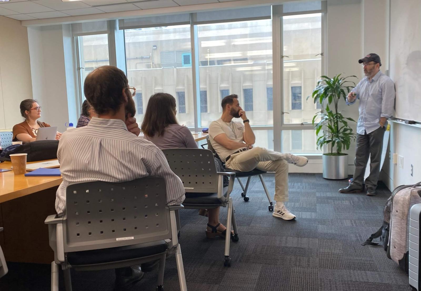 Dr. Adam Cohen stands by a whiteboard and speaks to a group of seated CASJE fellows, mentors, and staff.