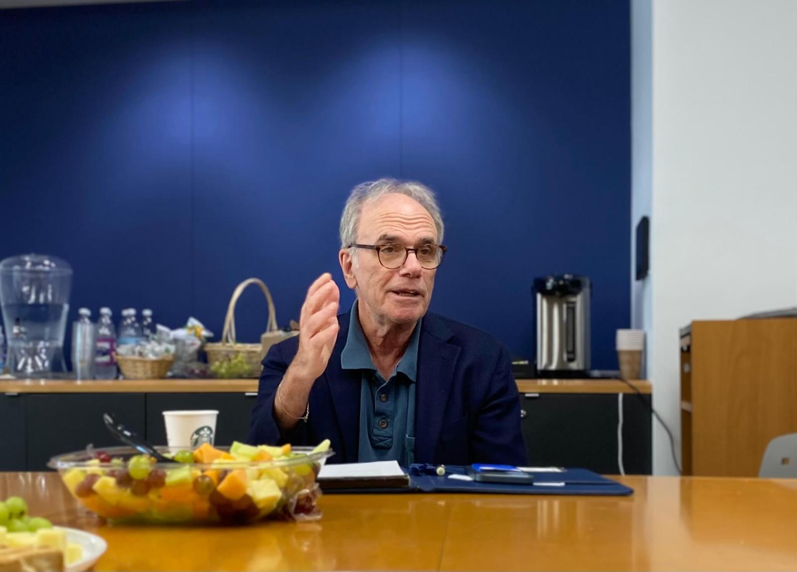 Dean Michael Feuer speaks in front of a blue background. He is seated at a brown table. A plate of fruit and a coffee cup are in front of him.