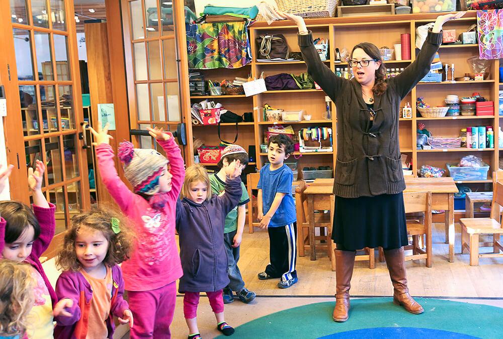 preschool teacher stands with arms raised high as preschool students by her follow her example