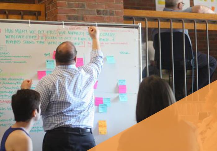 professional male writes on large white board with post-it notes scattered across it, small group watches
