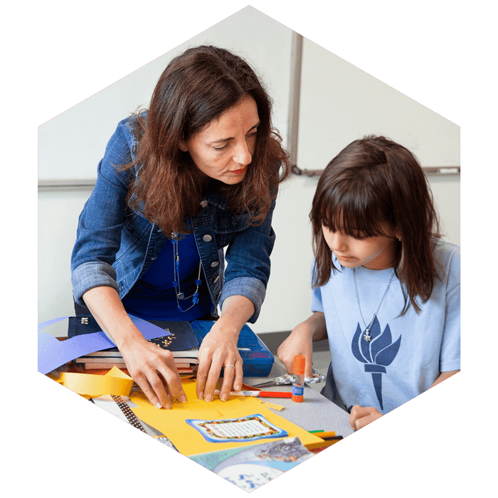Jewish Day School teacher assists elementary aged student with craft (image is hexagon shaped)