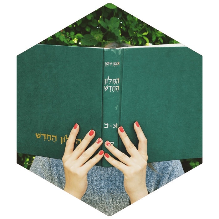girl holding Hebrew book with green cover open in front of her face (image is hexagon shaped)