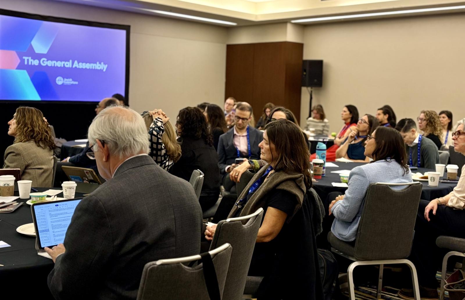Room of individuals facing the front. Screen reads JFNA General Assembly.
