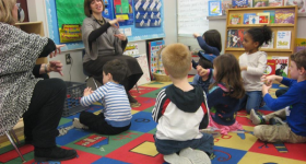 Children sit on a brightly colored carpet. A teacher sits in front of them.